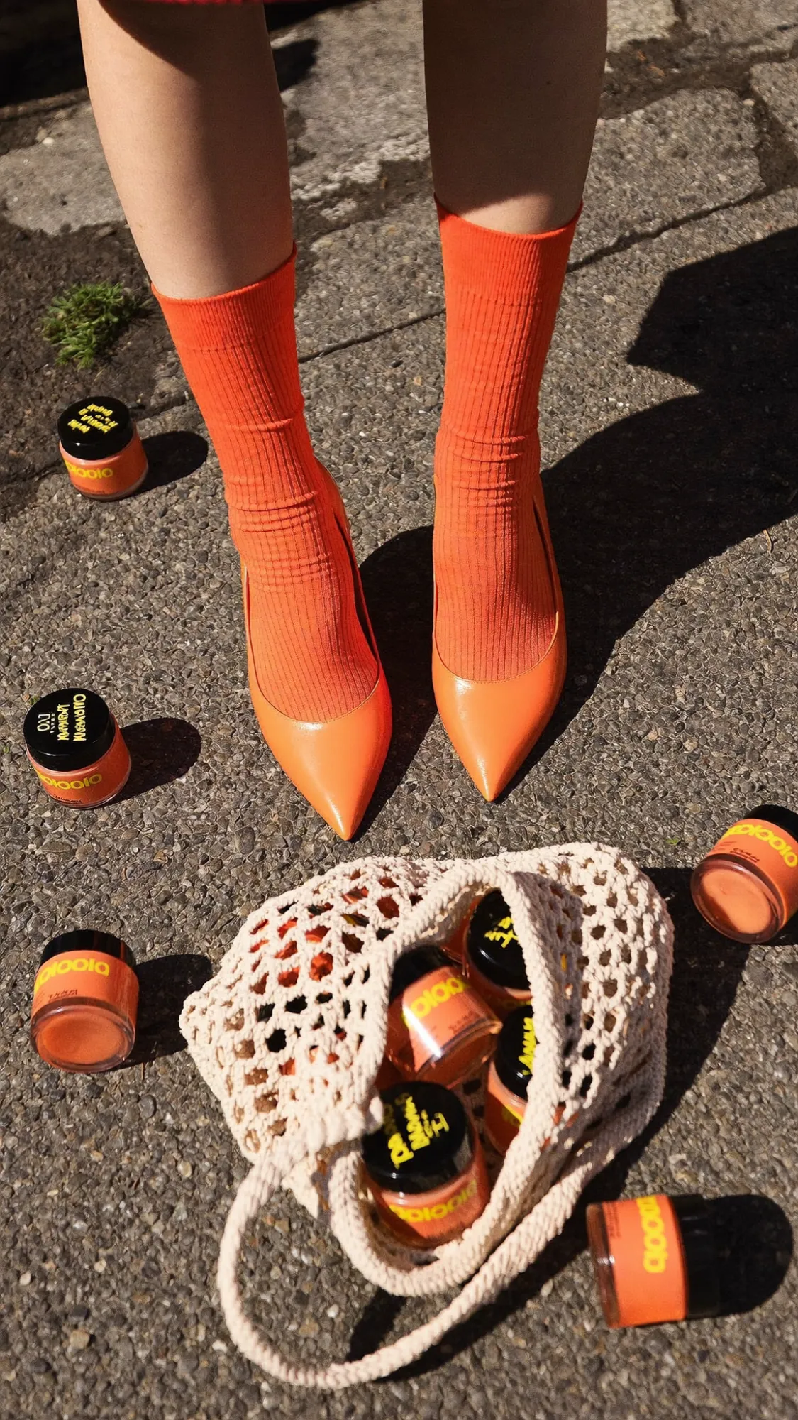 Orange ribbed socks and pointed heels on sunlit asphalt, with orange jars spilling from a crochet bag (concept)